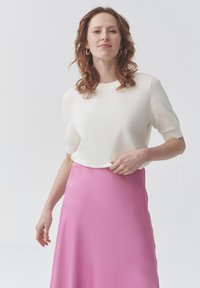 Bílý krátký svetr s krátkým rukávem a kulatým výstřihem v kombinaci s volnou růžovou sukní. Materiál vypadá měkce a hladce, s minimalistickým designem.