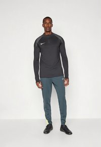 Čierne športové tričko s dlhými rukávmi, polovičný zips a sivé detailné pruhy na ramenách, v kombinácii s tyrkysovými teplákmi so žltými detailmi a čiernymi teniskami.