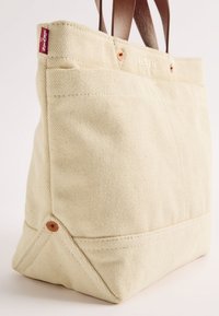 Beige canvas totebag met bruine leren handvatten, zichtbare Levi's logo tag en koperen klinknagel details op een plain witte achtergrond.