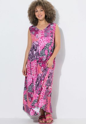 Mujer con cabello rizado que lleva un vestido maxi sin mangas con parche de paisley rosa y sandalias rosas a juego, posando contra un fondo blanco.