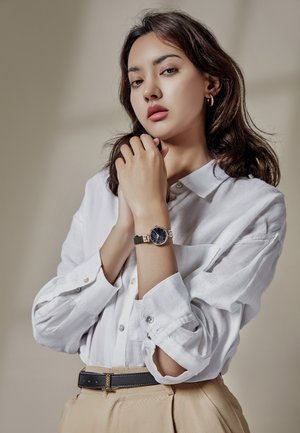 Femme aux longs cheveux noirs, portant une chemise blanche et un pantalon beige, avec une montre noire au poignet, posant avec les mains près de son visage sur un fond neutre.
