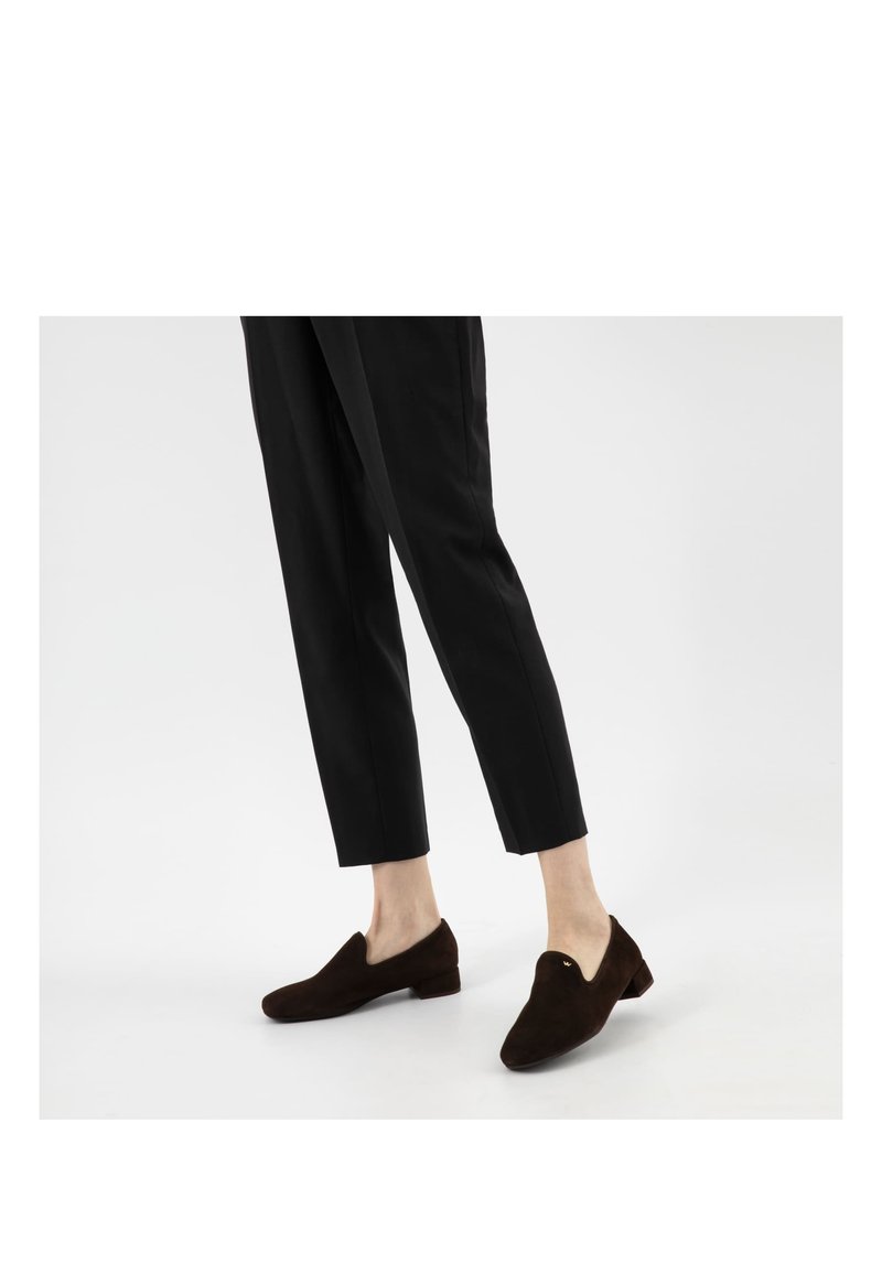 Osoba ubrana w krótkie czarne spodnie i ciemnobrązowe zamszowe mokasyny stojąca na jednolitym białym tle.