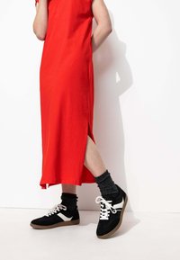 Robe rouge à mi-longueur avec des manches courtes et des fentes sur les côtés, associée à des baskets noires avec des accents blancs et des chaussettes grises texturées.