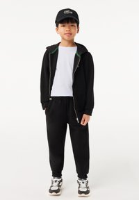 Schwarze Zip-Hoodie mit grünen Akzenten, weißes T-Shirt, schwarze Jogginghose und schwarze Mütze mit Logo; graue und weiße Sneaker runden den Look ab.