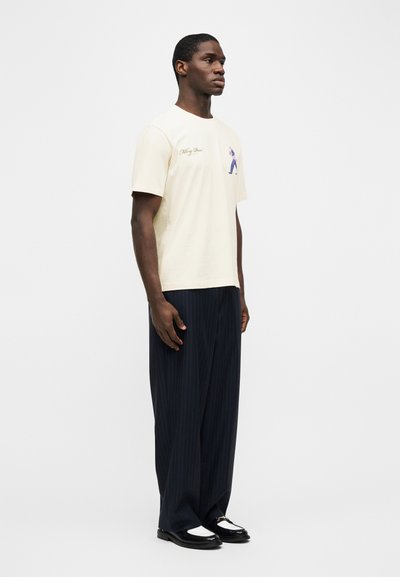 Άνδρας που φοράει κρεμ γραφικό T-shirt, σκούρο παντελόνι φαρδιά ρίγα, και μαύρα γυαλιστερά παπούτσια, στέκεται μπροστά σε απλό λευκό φόντο.