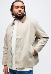 Άνδρας με πλεξούδες, που φοράει μπεζ σακάκι με γιακά από κορντούρα, λευκό πουλόβερ και μπλε τζιν, κοιτάζοντας προς τα πλάγια με το ένα χέρι στην τσέπη.