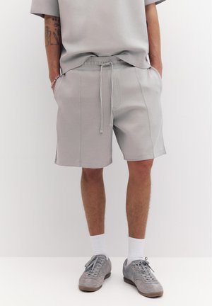 Mann, der hellgraue legere Shorts mit elastischem Bund, ein passendes lockeres T-Shirt, weiße Socken, graue Sneaker und ein silbernes Armband trägt.