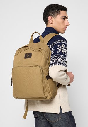 Mochila de tela kaki con forma rectangular, que cuenta con un bolsillo delantero con cremallera y detalles de cuero marrón. Correas ajustables para los hombros.