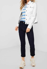 Chaqueta de mezclilla blanca, camisa de rayas azul y blanca, jeans oscuros y zapatillas crema. La chaqueta tiene botones y bolsillos; los jeans son ajustados y de longitud corta.