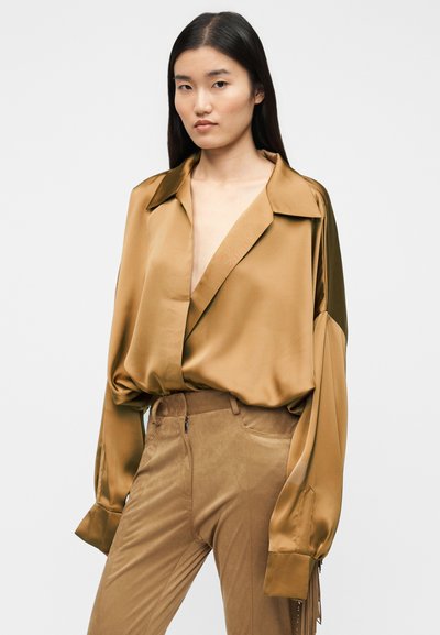 Μπλούζα από σατέν με μακριά μάνικα σε μπρονζέ χρώμα, με βαθύ V-λαιμόκοψη και φαρδύ γιακά. Συνδυασμένη με παντελόνι από καφέ σουέντ με ομαλή υφή.