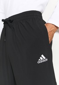 Schwarze Sporthose mit einem elastischen Bund und Kordelzug. Mit Seitentaschen und einem weißen Adidas-Logo am linken Bein. Glatte Textur.
