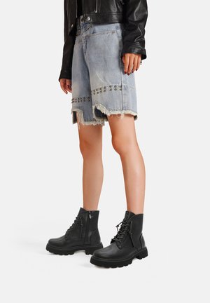 Stivaletti neri alla caviglia con finitura texture, lacci davanti e zip laterale. Abbinati a pantaloncini di jeans sfilacciati con borchie metalliche.