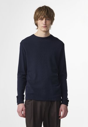 Young man with medium-length brown hair wearing a long-sleeve navy shirt and dark trousers, standing against a plain white background.