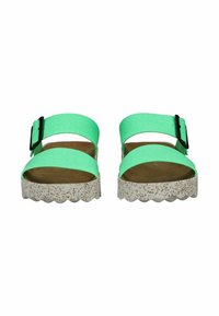 Cinghie di tessuto verde con una fibbia nera, soletta morbida marrone e una suola piatta bianca testurizzata con bordi ondulati.