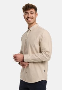 Camisa beige de botones con cuello, mangas largas y un sutil detalle de logo en el pecho. Fabricada con una tela suave y un corte ajustado.