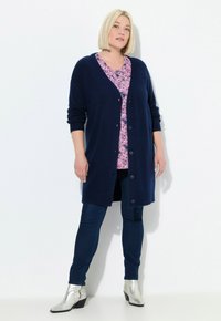 Marinblå stickad cardigan med v-ringning och knappstängning, bärs över en rosa blommig topp, matchad med mörka jeans och silverfärgade ankelstövlar.