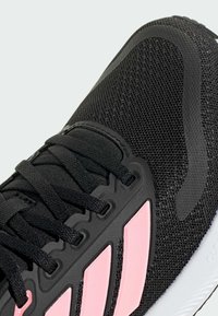 Chaussure de sport noire avec des accents roses, tige en mesh, semelle en caoutchouc texturée et fermeture à lacets. Design remarquable avec trois rayures sur le côté.