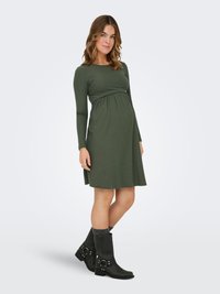 Raskaana oleva nainen seisoo vaaleaa taustaa vasten pukeutuneena pitkähihaiseen oliivinvihreään äitiysmekkoon ja mustiin puolivälikorkaisiin saappaisiin harmaiden sukkien kanssa.