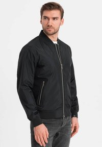 Veste bomber noire en nylon lisse avec fermeture éclair, poignets côtelés et deux poches zippées sur les côtés. Présente un design de col classique.