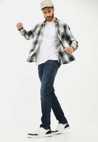 Chemise en flanelle à carreaux bleue et beige, T-shirt blanc, jeans foncés et baskets blanches avec accents marine. Homme en position debout.
