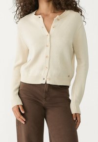 Sweter w kolorze kremowym, wykonany na drutach, z okrągłym dekoltem, długimi rękawami i zapięciem na guziki. Posiada ściągacze na mankietach i u dołu, wykonany z miękkiego materiału.