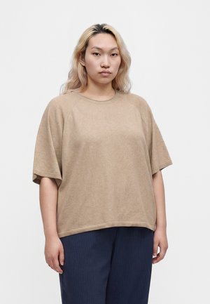 Ung person med langt lyst blondt hår iført en løs beige kortærmet top og mørkeblå bukser, der står mod en ensfarvet hvid baggrund.