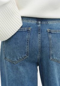 Denimjeans i en blekt blå färg, med en klassisk femfickorsdesign och framträdande bakfickor. Ribbade vita tröja synlig upptill.