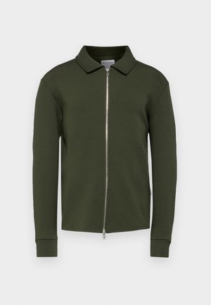 Veste zippée verte en tissu doux. Elle présente un col plié, des manches longues et des poignets côtelés, avec une fermeture éclair centrale en métal.