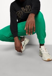 Schwarzer Langarm-Sweatshirt mit goldener Grafik, grüne Sweatpants und weiße Sneakers mit grauen und grünen Akzenten. Person in der Hocke.