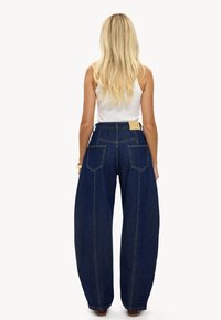 Jeans de pierna ancha y talle alto en denim oscuro, con acabado limpio, dos bolsillos traseros y detalles sutiles de costura, combinados con una camiseta blanca.