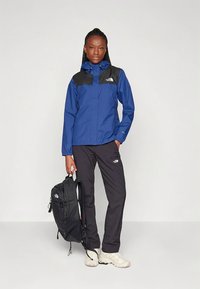 Casaco impermeável azul com detalhes pretos nos ombros, combinado com calças pretas, segurando uma mochila preta e usando sapatos de cor clara.