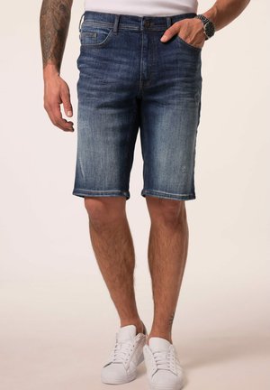 Mann trägt dunkelblaue Denim-Shorts und weiße Sneakers, steht mit einer Hand in der Tasche und sichtbar tätowiertem Unterarm.