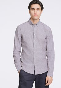 Camisa de manga larga, con botones, en tela gris claro con una textura sutil. Presenta un cuello clásico y botones blancos. Combinada con pantalones oscuros.
