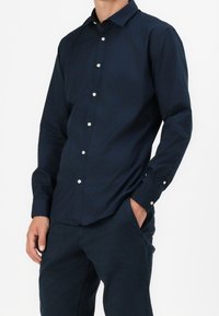Camicia button-up blu navy con colletto, dotata di bottoni bianchi, maniche lunghe e una texture liscia, indossata con pantaloni blu navy coordinati.