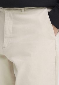 Bēša kokvilnas bikses ar gludu tekstūru, kurās ir priekšējā kabata un jostas cilpas, kopā ar melnu garroku topu.