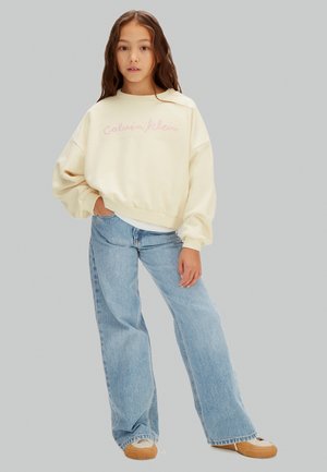 Niña de pie con la mano en el bolsillo, vistiendo un suéter crema de Calvin Klein, jeans acampanados azul claro y zapatillas beige sobre un fondo gris.