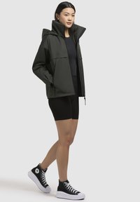 Giacca impermeabile verde scuro con colletto alto, tasche frontali e cerniere, abbinata a pantaloni corti neri da bici e sneaker nere con plateau.