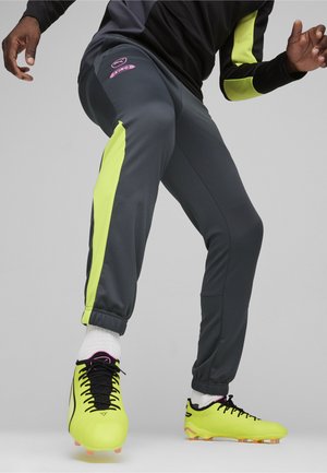 Grijze sportieve herenbroek met neongele zijpanelen, elastische boorden en een paarse logo. Gecombineerd met felgele voetbalschoenen.