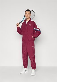 Maroon sportska jakna i hlače s bijelim i plavim akcentima. Ima patentni zatvarač, elastične manžete i džepove. Nosite s bijelim sportskim cipelama.