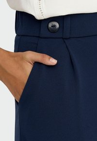 Marineblå bukser med en glat tekstur, med knaplukning og sidelommer, med en hånd hvilende i lommen.