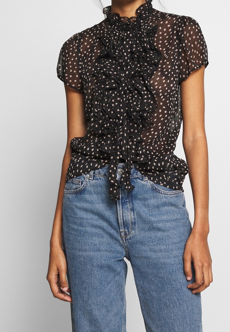 Femme portant un chemisier noir transparent à manches courtes avec des pois blancs et des volants, associé à un jean bleu taille haute.