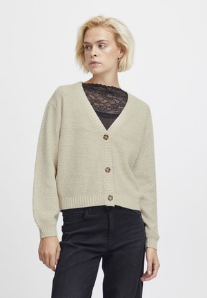 Beige neulecardigan, jossa on V-pääntie, viisi ruskeaa nappia ja ribbireunat, päällystetty mustan pitsitopin yli ja yhdistetty mustiin housuihin.