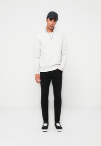 Un jeune homme portant un pull gris à fermeture éclair quart, un pantalon noir, une casquette noire et des baskets noires avec des lacets blancs, se tenant contre un fond blanc.