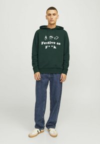 Felpa verde con un design frontale che recita "Festivo come F***", con icone di un albero, Babbo Natale e una bastoncino di zucchero. Abbinata a jeans blu e sneaker.