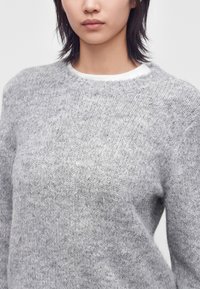 Femme aux cheveux raides et foncés portant un pull en maille doux et texturé gris clair sur un t-shirt blanc à col rond, présentée des épaules au milieu du buste.