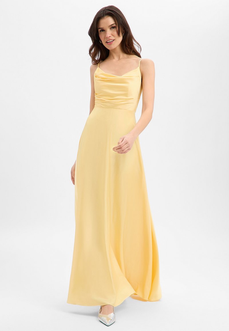 Femme portant une robe fluide en satin jaune à fines bretelles, souriante, posant devant un fond blanc uni.