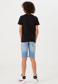 T-shirt nera in cotone, pantaloni corti in denim blu e sneakers bianche con accenti neri e rossi. Design visibile sul retro, outfit estivo casual.