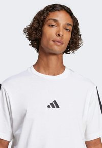 Biela bavlnená tričko s čiernym logom Adidas na hrudi, s čiernymi akcentmi na rukávoch a kulatým výstrihom.