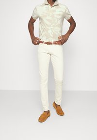 Světle béžové polo tričko s kamuflážním vzorem, krémové upnuté kalhoty a hnědý pletený pásek. Semišové mokasíny v teplém hnědém odstínu. Jednoduchý, elegantní vzhled.