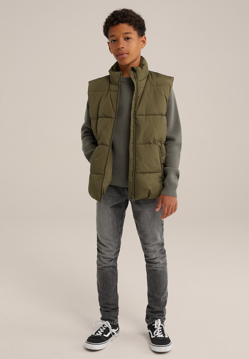 Olivgrön pufferväst med hög krage, bärs över en grå tröja. Matchad med smala grå jeans och svarta sneakers med vita sulor.